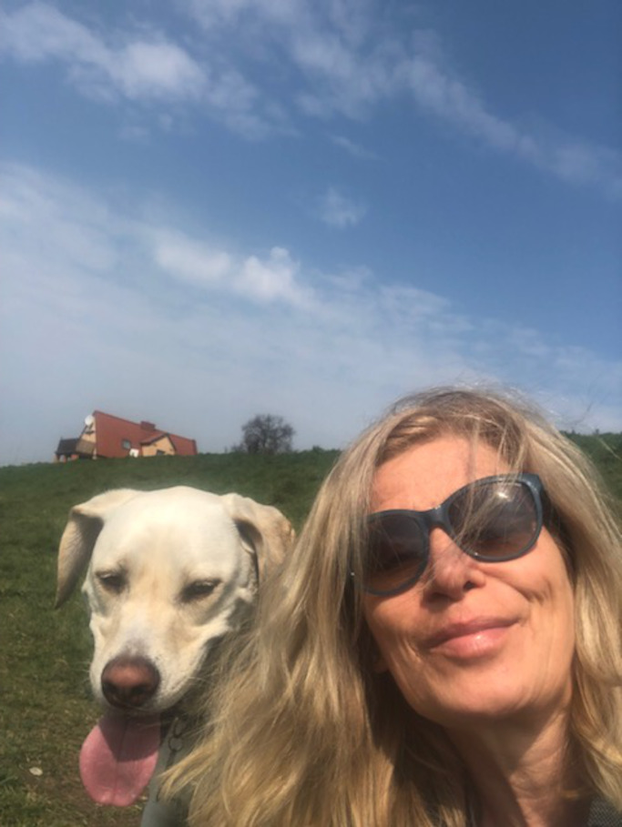 Uśmiechnięta blondynka z okularami przeciwsłonecznymi na twarzy, obok niej biały pies rasy labrador, w tle zielona łąka i błękitne niebo.