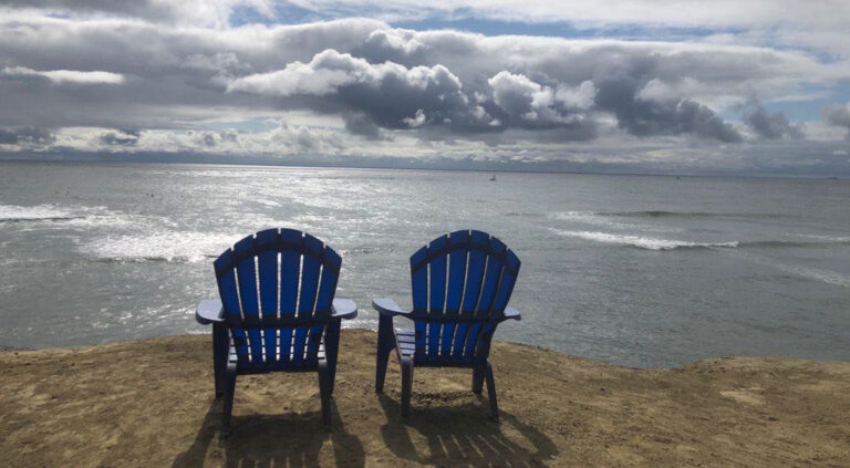 Na pustej plaży stoją dwa niebieskie krzesła zwrócone ku morzu. Na morzu są nieliczne fale. Słońce przebija się zza chmur i widać elementy niebieskiego nieba.