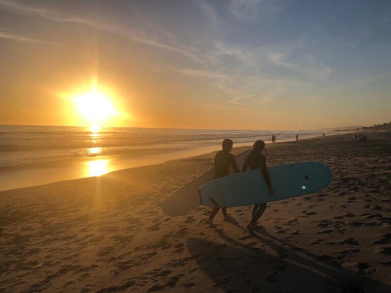 Kobieta i mężczyzna idą po plaży trzymając deskę do surfingu, w tle zachodzące słońce